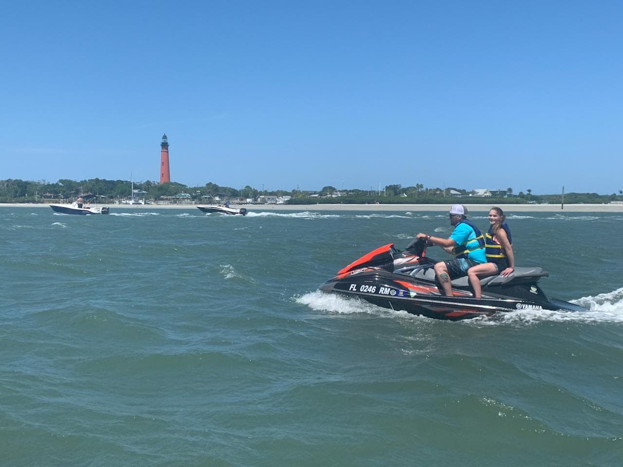 A couple riding tandem across the inlet with the lighthouse and the beach on the far shore.